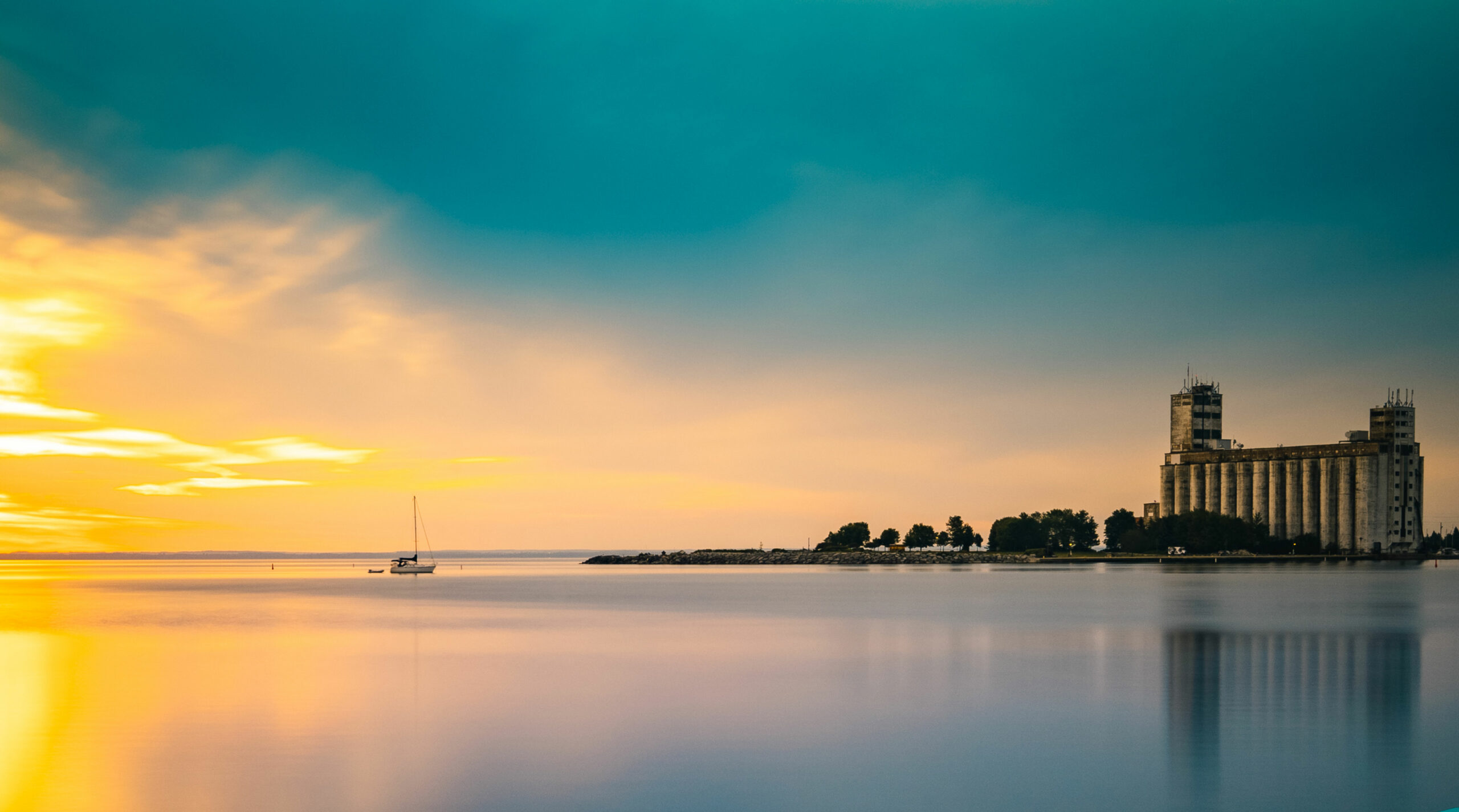 A calm waterfront scene in Collingwood at sunset with a solitary sailboat and a large building on the right under a pastel sky.