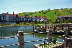 A picturesque lakeside resort with Tudor-style architecture set against a green hill under a clear blue sky.